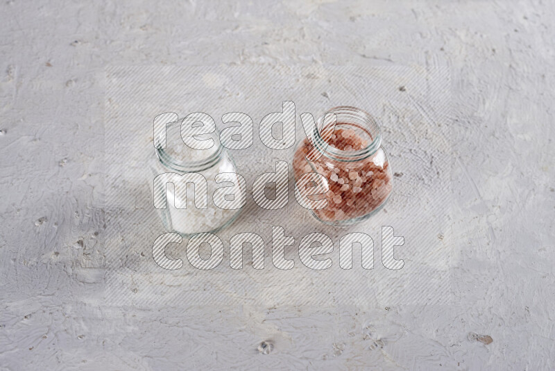 2 glass jars one is filled with coarse himalayan salt and the other with coarse sea salt on white background