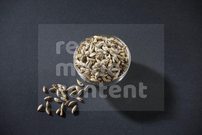 A glass bowl full of cardamom and more seeds spreaded beneath the bowl on black flooring