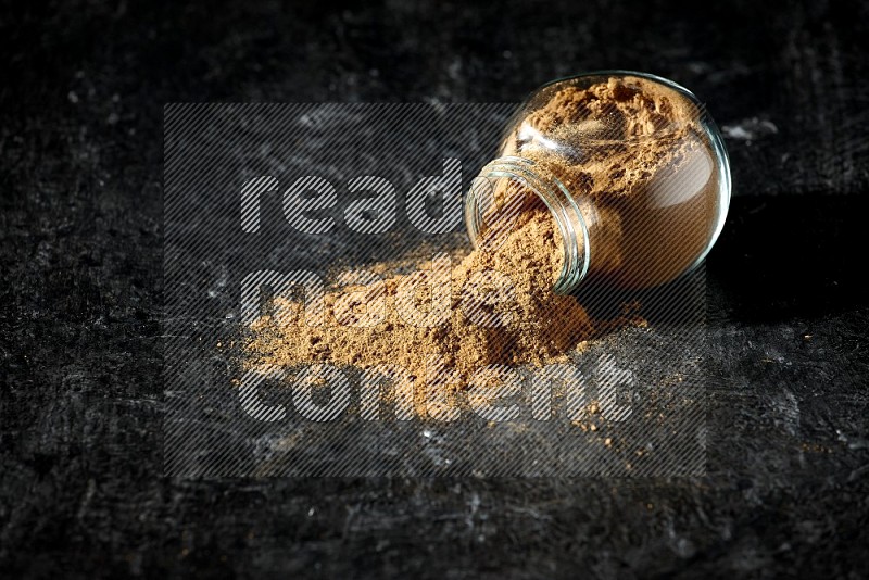 A flipped glass spice jar full of allspice powder and powder spilled out of it on a textured black flooring