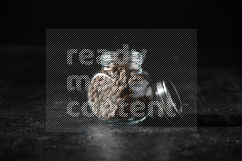 A glass spice jar full of allspice whole balls on a textured black flooring