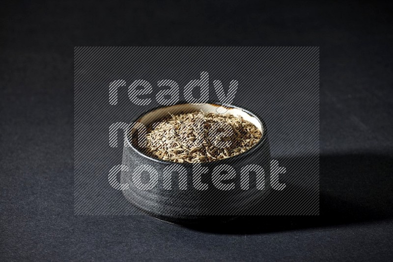 A black pottery bowl full of cumin seeds on a black flooring