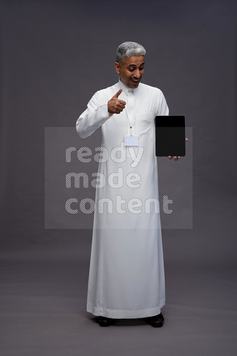 Saudi man wearing thob with neck strap employee badge standing showing tablet to camera on gray background