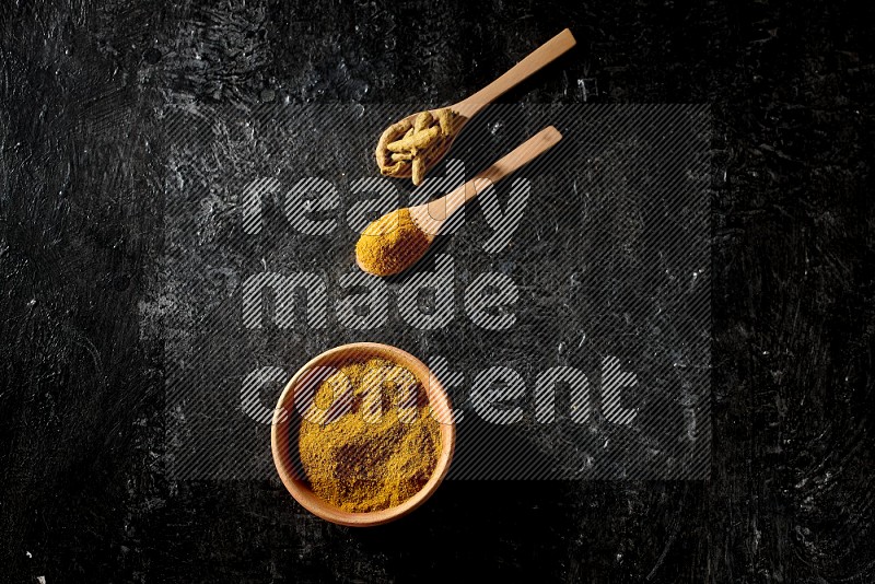 A wooden bowl full of turmeric powder and 2 wooden spoons full of dried turmeric finger and turmeric powder on textured black flooring