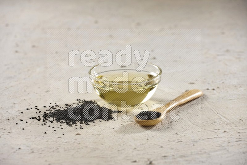A glass bowl full of black seeds oil and wooden spoon full of black seeds with seeds spread on a textured white flooring