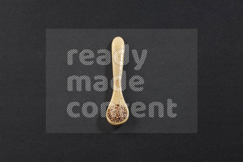 A wooden spoon full of mustard seeds on a black flooring