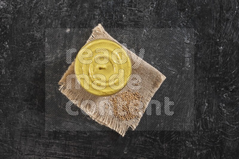 A glass bowl full of mustard paste set on a burlap piece with some mustard seeds on a textured black flooring