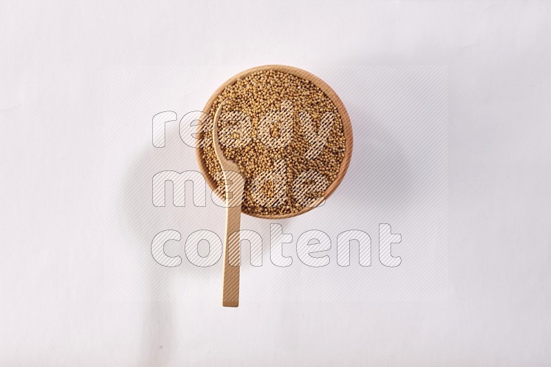 A wooden bowl full of mustard seeds with a wooden spoon on the seed on a white flooring