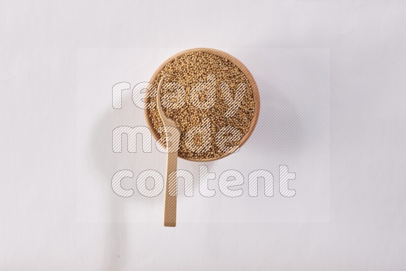A wooden bowl full of mustard seeds with a wooden spoon on the seed on a white flooring