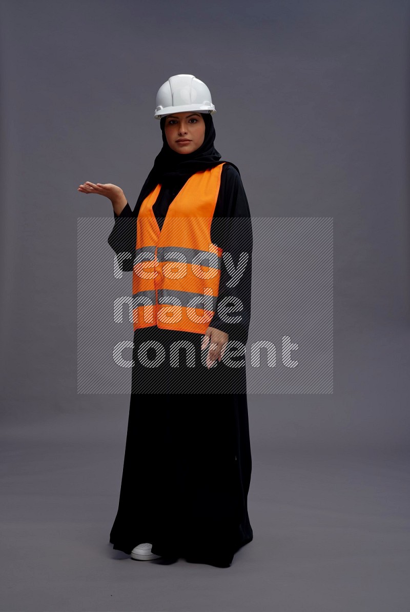 Saudi woman wearing Abaya with engineer vest standing interacting with the camera on gray background