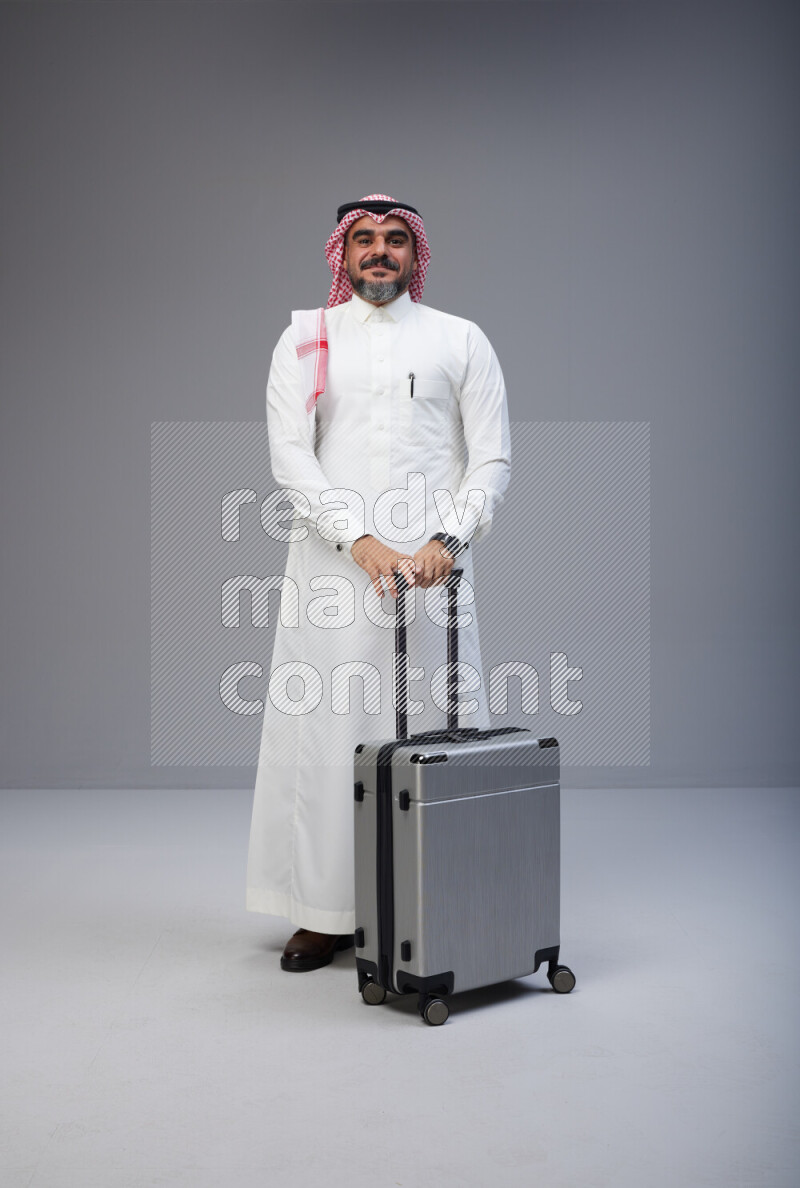 Saudi man wearing Thob and red Shomag standing holding Travel bag on Gray background