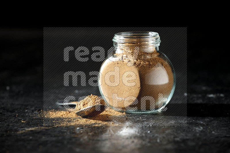 A glass spice jar and metal spoon full of allspice powder on a textured black flooring