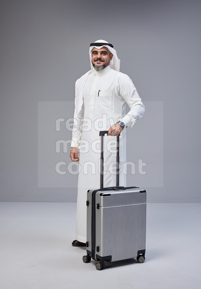 Saudi man wearing Thob and white Shomag standing holding Travel bag on Gray background