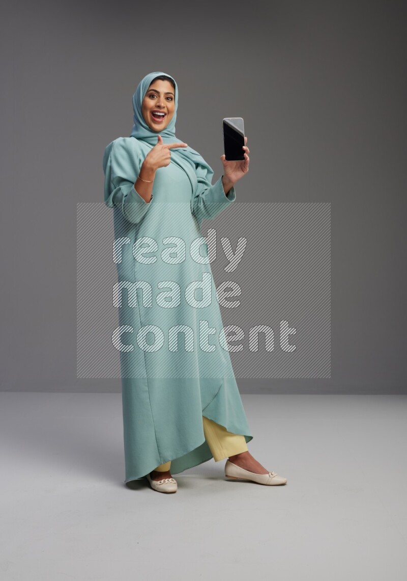 Saudi Woman wearing Abaya standing showing phone to camera on Gray background