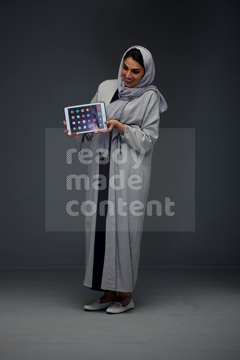 A Saudi woman wearing a light gray Abaya and head scarf standing and showing the phone's screen eye level on a grey background