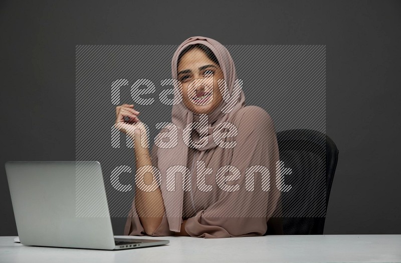 A Saudi woman Setting on her desk on a Gray Background wearing Brown Abaya with Hijab
