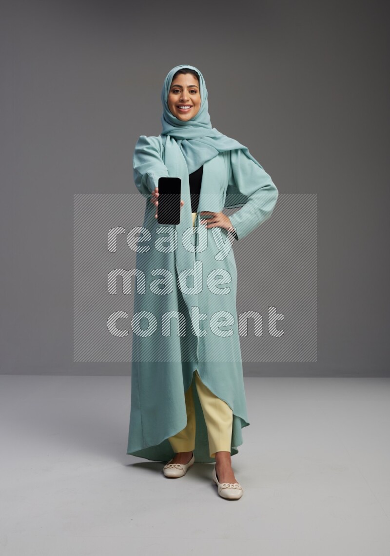 Saudi Woman wearing Abaya standing showing phone to camera on Gray background