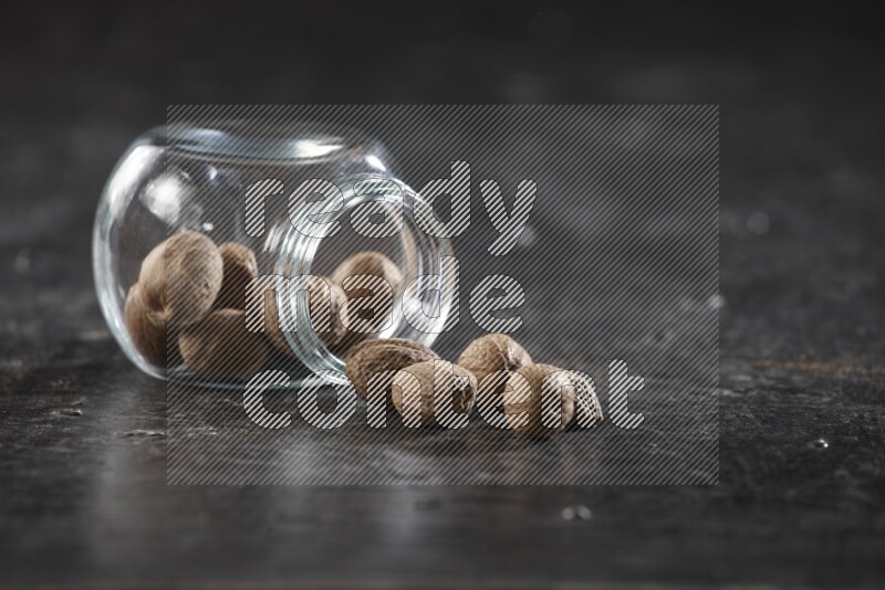 A glass spice jar full of whole nutmeg seeds flipped and the seeds spread out on a textured black flooring