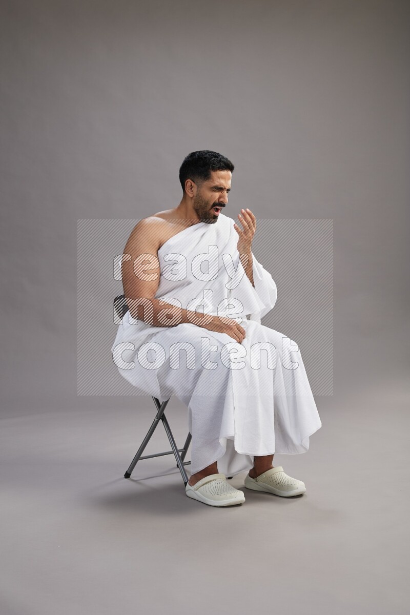 A man wearing Ehram sitting on chair showing how to sneeze on gray background