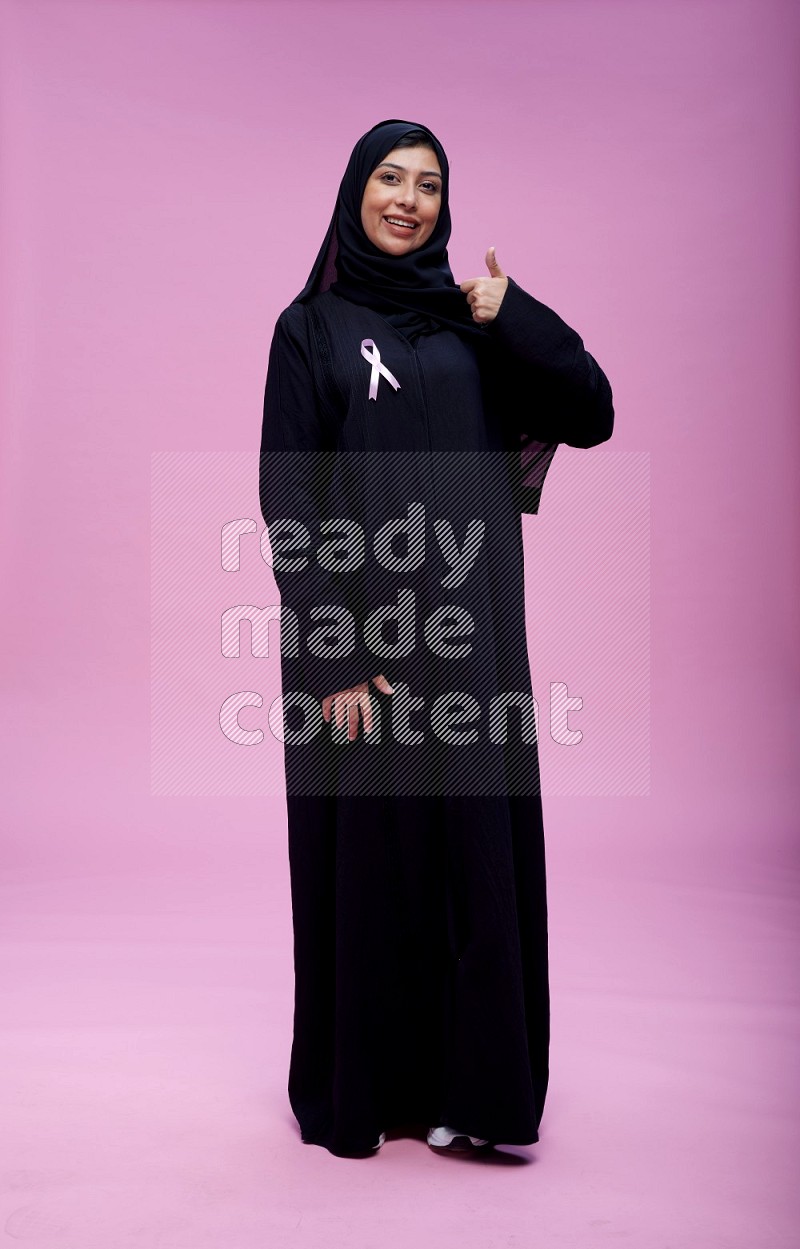 Saudi woman wearing pink ribbon on Abaya standing interacting with the camera on pink background