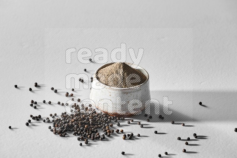 A white ceramic bowl full of black pepper powder and black pepper beads spread on white flooring