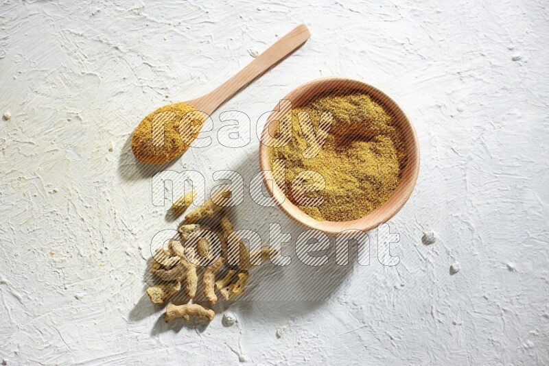 A wooden bowl and wooden spoon full of turmeric powder with dried turmeric fingers on textured white flooring