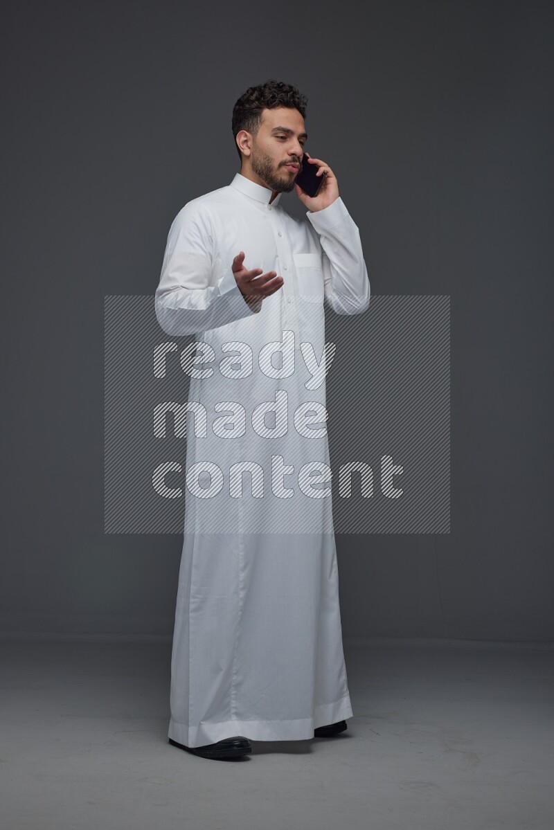 A Saudi man wearing Thobe and talking in the phone while standing and making different poses eye level on a gray background