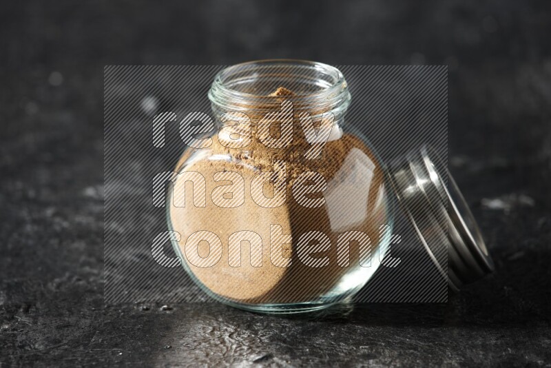 A glass spice jar full of allspice powder on a textured black flooring