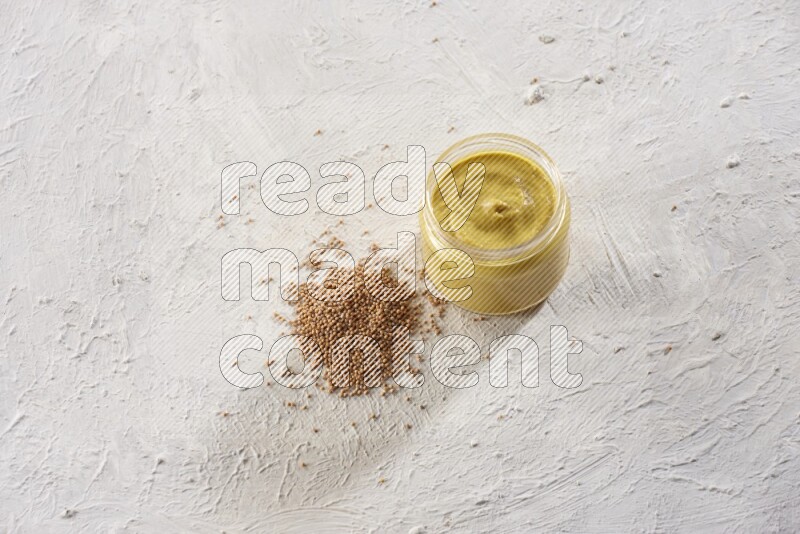 A glass jar full of mustard paste with mustard seeds spread next to it on a textured white flooring