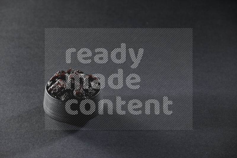 A black pottery bowl full of dried plums on a black background in different angles