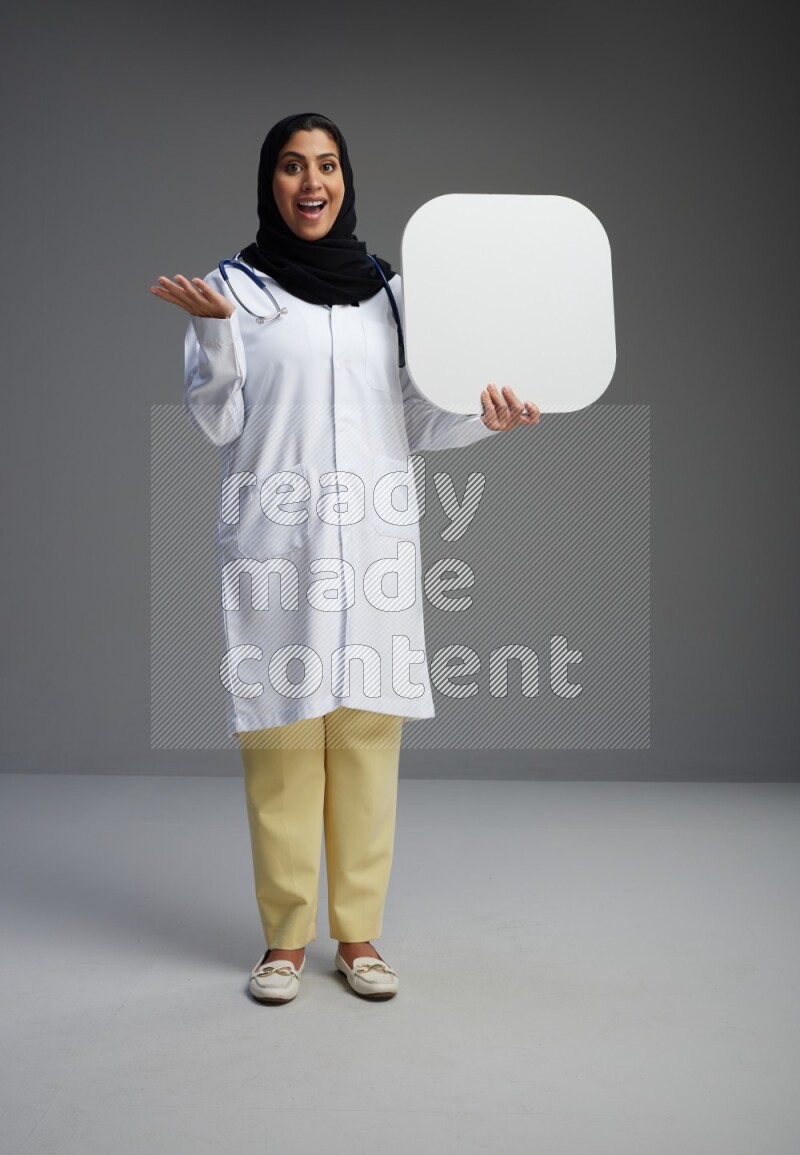 Saudi woman wearing lab coat with stethoscope standing holding social media sign on Gray background