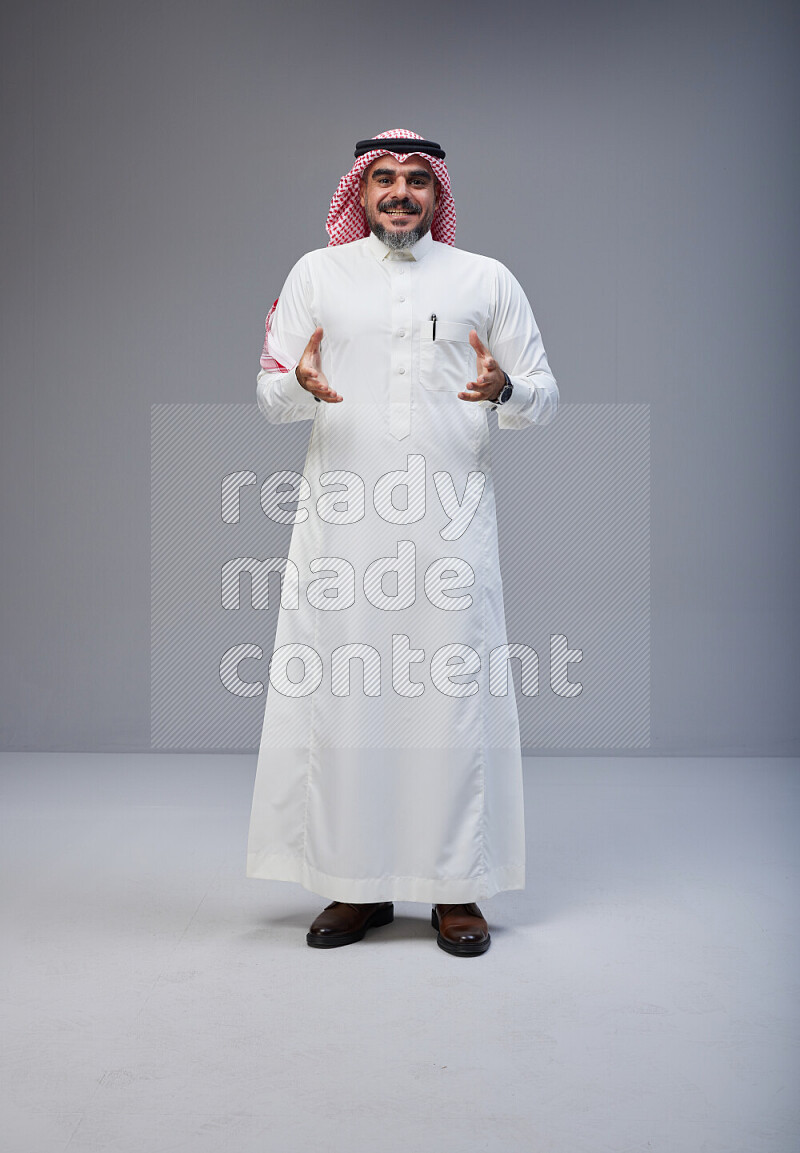 Saudi man Wearing Thob and red white Shomag standing interacting with the camera on Gray background