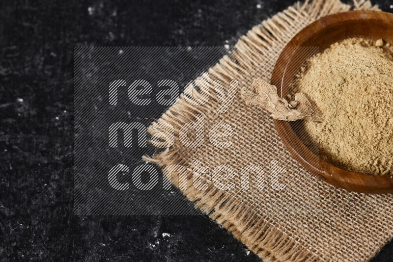 A wooden bowl full of ground ginger powder with a wooden spoon on it all on a burlap fabric on black background