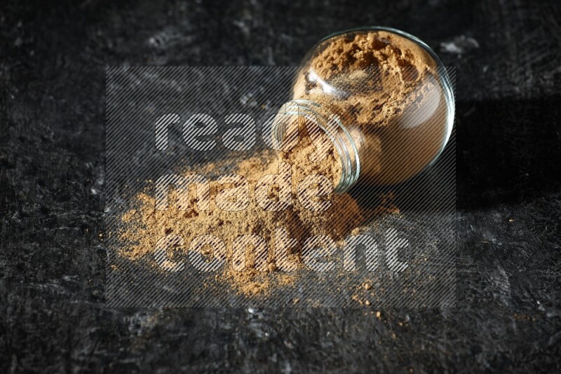 A flipped glass spice jar full of allspice powder and powder spilled out of it on a textured black flooring