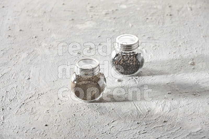 2 glass spice jars full of black pepper and black pepper powder on textured white flooring