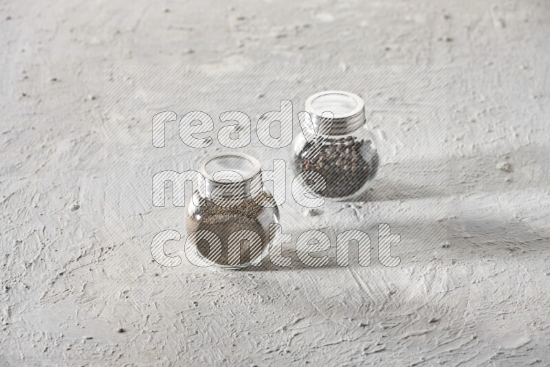 2 glass spice jars full of black pepper and black pepper powder on textured white flooring