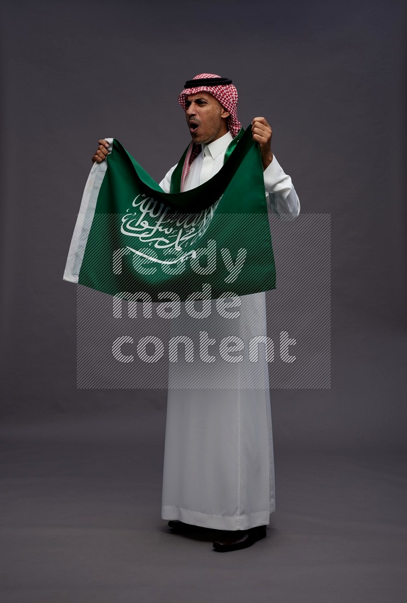 Saudi man wearing thob and shomag standing holding Saudi flag on gray background