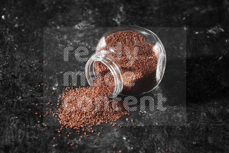 A glass spice jar full of garden cress seeds flipped and seeds spread out a textured black flooring