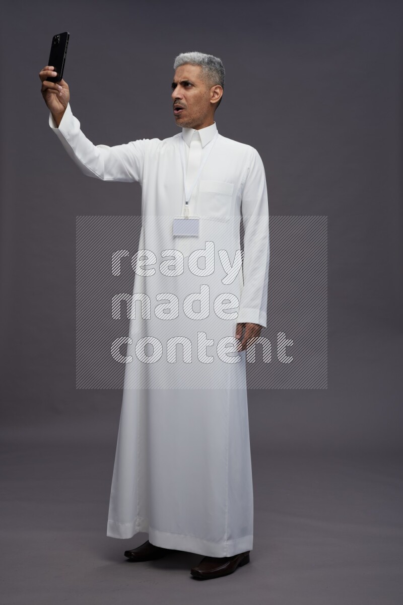 Saudi man wearing thob with neck strap employee badge standing taking selife on gray background