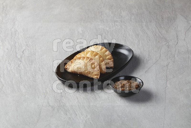Three fried sambosas in an oval shaped black plate and a sauce in a black round ramekin on a gray background