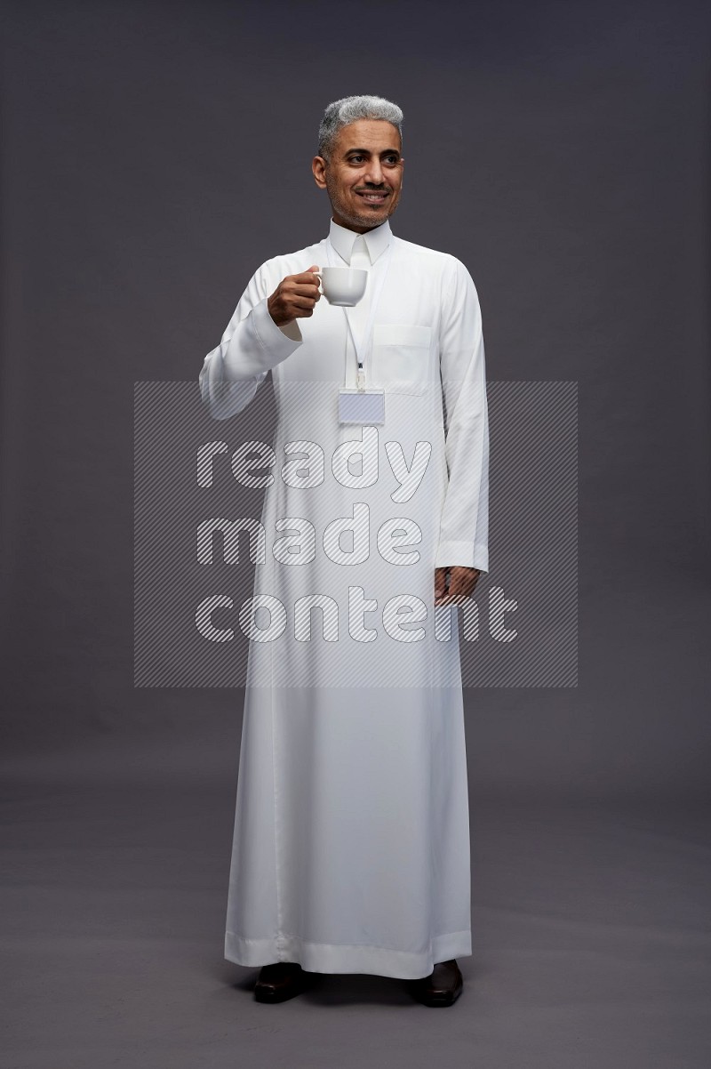 Saudi man wearing thob with neck strap employee badge standing holding mug on gray background