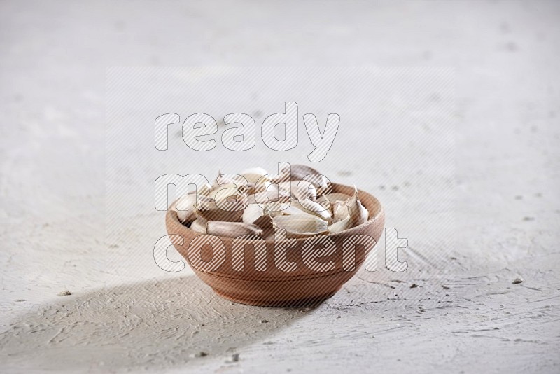 A wooden bowl full of garlic cloves on a textured white flooring in different angles