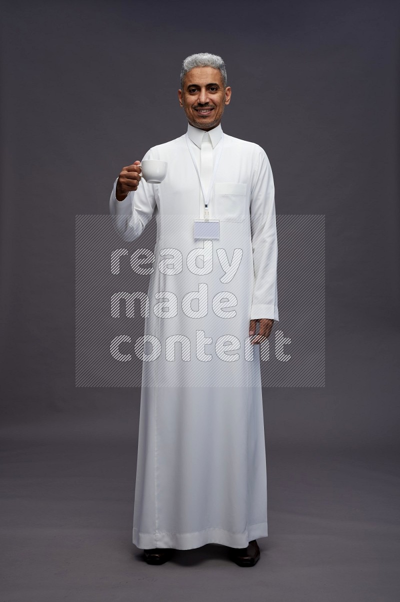 Saudi man wearing thob with neck strap employee badge standing holding mug on gray background