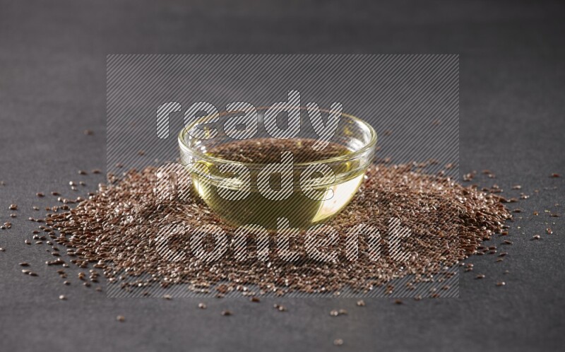A glass bowl full of flaxseeds oil surrounded by the seeds on a black flooring