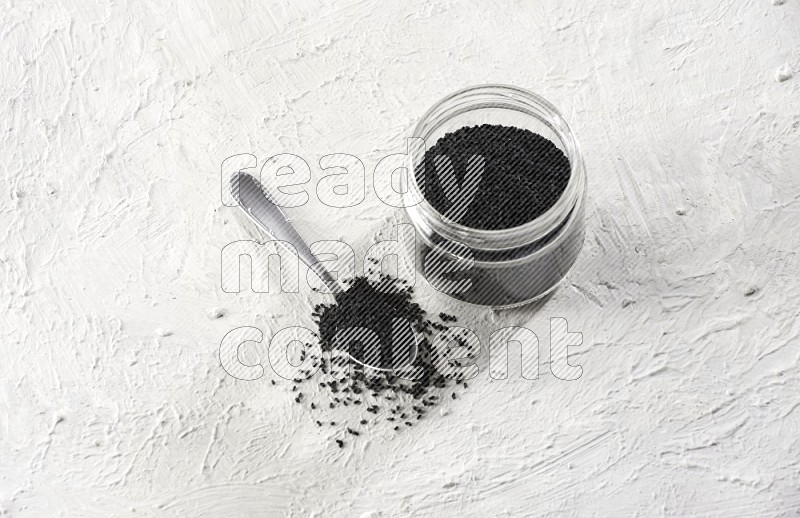A glass jar and a metal spoon full of black seeds on a textured white flooring in different angles
