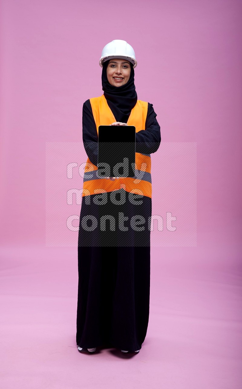 Saudi woman wearing Abaya with engineer vest and helmet standing showing tablet to camera on pink background