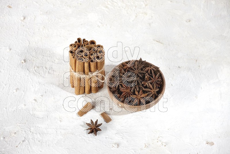 A stacked and bounded cinnamon sticks and a wooden bowl full of star anise on a white background