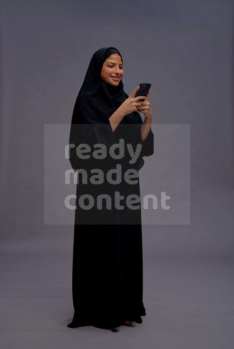 Saudi woman wearing Abaya standing texting on phone on gray background