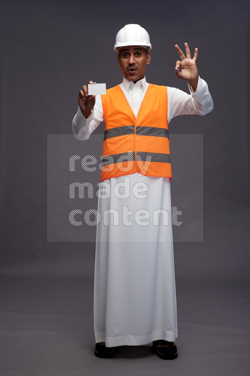 Saudi man wearing thob with engineer vest standing holding ATM card on gray background