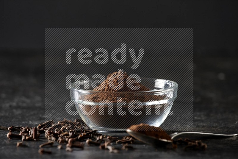 A glass bowl and a metal spoon full of cloves powder with gloves grains beside them on a textured black flooring