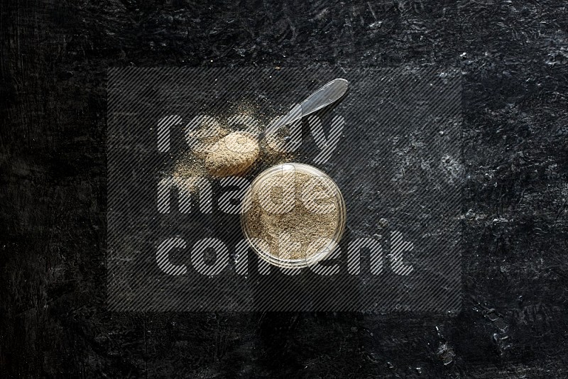 A glass jar and metal spoon full of cardamom powder on textured black flooring